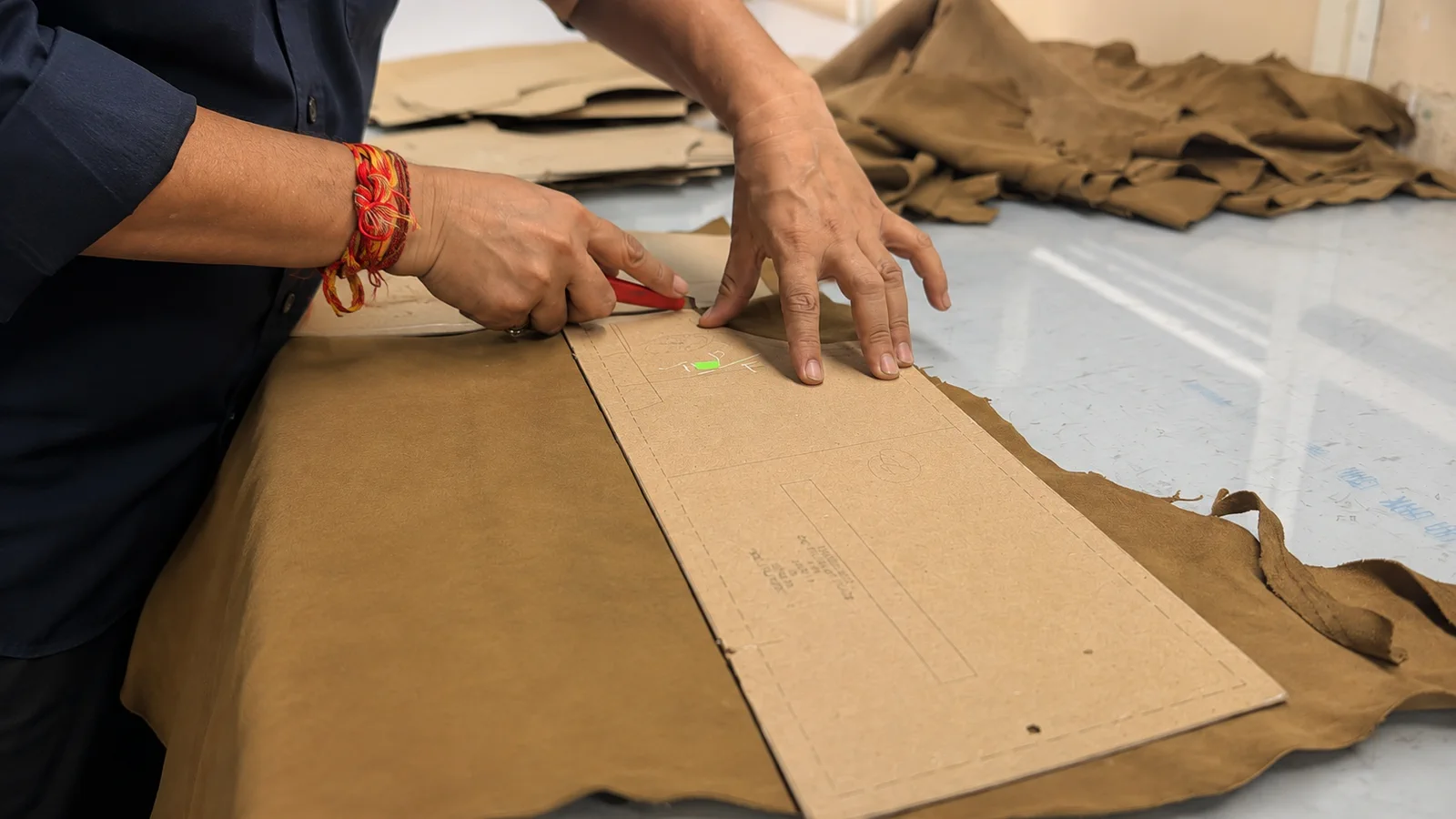 Hands cutting a leather skin around a paper pattern with a knife.